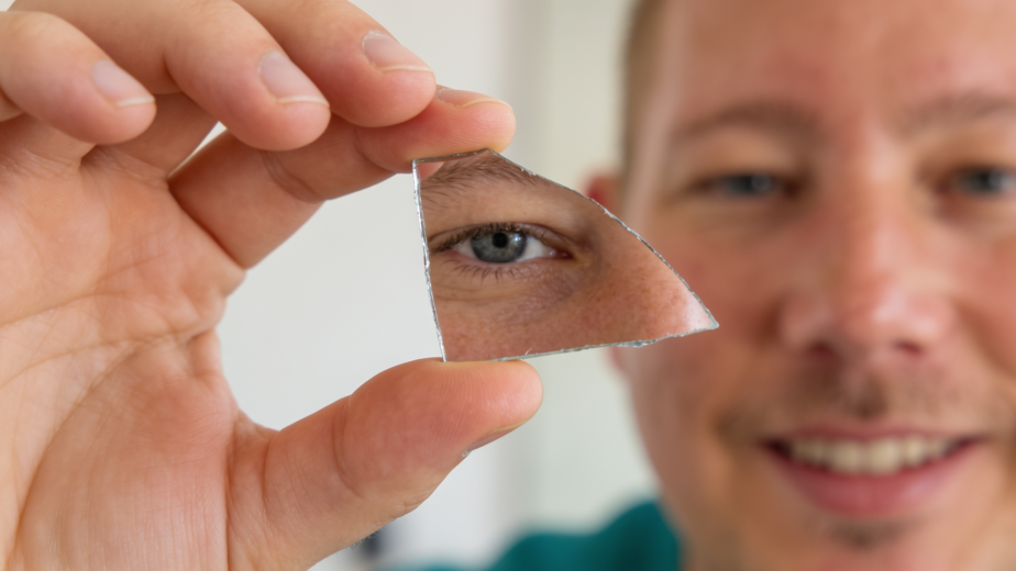Man holding a small mirror shard, catching a partial reflection of his own eye, representing the practice of shadow work and self-reflection