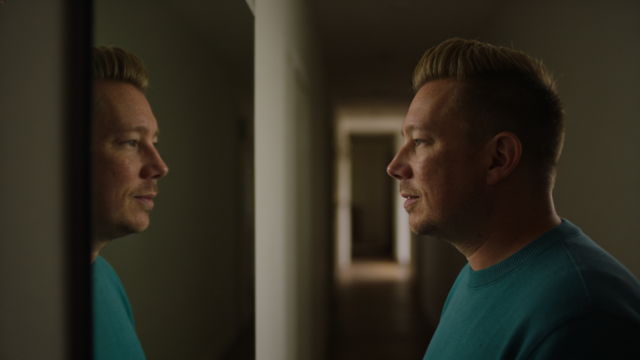 Man standing in a hallway looking at his reflection in a mirror with a quiet, contemplative expression, representing the process of recognising and understanding repeating patterns in spiritual growth